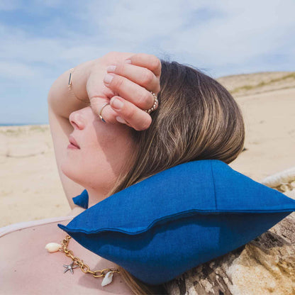 Femme allongée sur le sable, adossée à un bois flotté, avec coussin cervical Cosy Club bleu derrière le cou pour éviter les tensions à la nuque